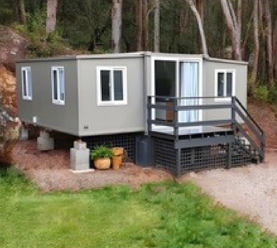Grey Expandable Home raised on stilts with a railed deck in a forest.