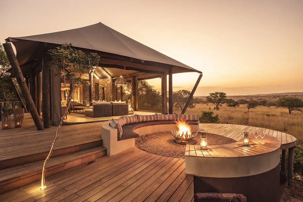 Curved bench with striped cushions around a fire pit on the deck of a canvas-roofed Safari Tent suite at sunset.
